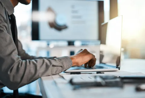شخص يرتدي قميصًا مخططًا يعمل على كمبيوتر محمول في مكتب حديث، مع ظهور شاشات تعرض بيانات، مما يعكس بيئة العمل في مجال الذكاء الاصطناعي.