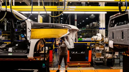 عامل في مصنع سيارات يقوم بتجميع هيكل سيارة، مع وجود معدات وأجزاء سيارات في الخلفية، مما يعكس تأثير الرسوم الجمركية على الصناعة.