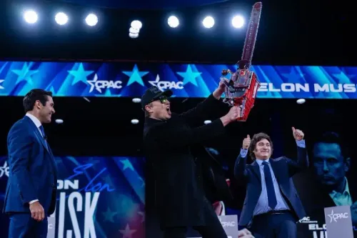 إيلون ماسك يحمل منشارًا كهربائيًا على المسرح في مؤتمر CPAC، مع جمهور متحمس، في مشهد يرمز لتحدي البيروقراطية الفيدرالية.