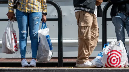 أشخاص ينتظرون في محطة مع أكياس تسوق تحمل شعارات متاجر، مما يعكس تراجع معنويات المستهلكين في ظل المخاوف الاقتصادية.