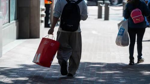 رجل يحمل حقيبة تسوق حمراء في شارع مزدحم، بينما يسير بجوار امرأة تحمل حقيبة وردية، مما يعكس الأجواء الاقتصادية الحالية.