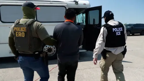 احتجاز مهاجر غير شرعي من قبل ضباط إنفاذ قوانين الهجرة، مع وجود حافلة للترحيل في الخلفية، يعكس سياسة الترحيل الحالية في الولايات المتحدة.