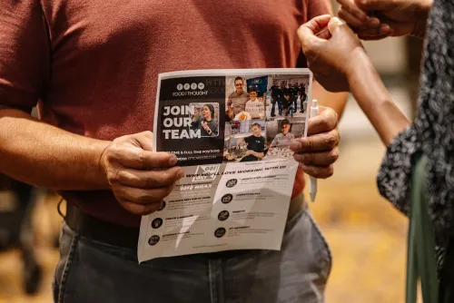 شخص يحمل منشورًا عن فرص العمل، مع صور لموظفين، في سياق مناقشة حول تحديات سوق العمل في الاقتصاد الأمريكي.