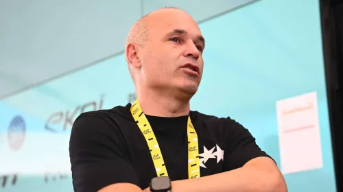 أندريس إنييستا يرتدي قميصاً أسود مع شارة صفراء حول عنقه، يظهر وهو يتحدث في حدث رياضي، مع خلفية شاشة عرض تحمل شعارات.