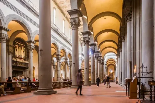 مشهد داخلي لدير سانتو سبيريتو في فلورنسا، يظهر أعمدة رائعة وأشخاص يتجولون، مع التركيز على تاريخ الدير ومعركته ضد خطط تحويله إلى دار مسنين فاخرة.