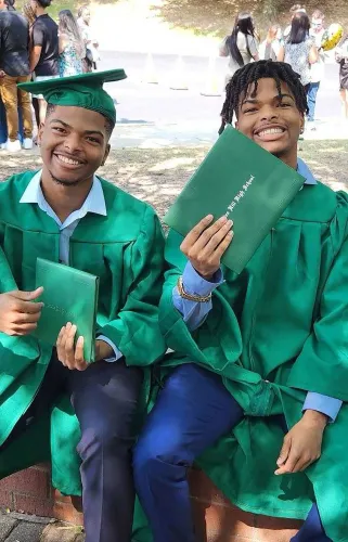 شقيقان توأمان يرتديان قبعات التخرج والعباءات الخضراء، يجلسان معاً مبتسمين ويحتفظان بشهادتيهما، في أجواء احتفالية.