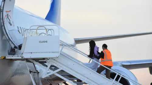 صورة لمهاجرين يتجهون إلى طائرة مع موظف يرتدي سترة برتقالية، تعكس إجراءات الترحيل القسري في ظل قوانين الهجرة.