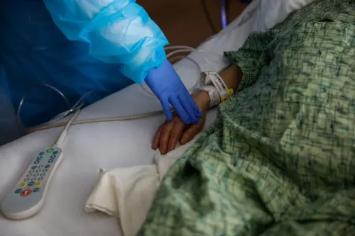 يد تشير يد مهنية ترتدي قفازًا أزرق إلى يد مريض مستلقٍ على سرير مستشفى، مما يعكس التحديات التي تواجه النساء في القوى العاملة خلال فترة الجائحة.