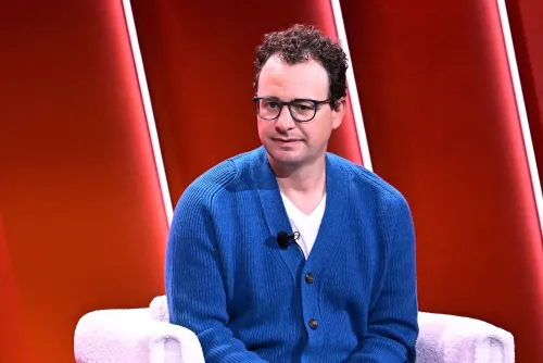 رجل يرتدي سترة زرقاء يجلس على كرسي أبيض، مع خلفية حمراء، أثناء حديثه عن سياسة السلامة الجديدة لشركة أنثروبيك في مجال الذكاء الاصطناعي.