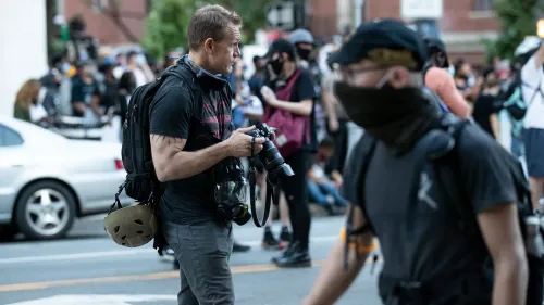 مصور محترف يحمل كاميرا ويتجول بين حشد من المتظاهرين، يعكس التوترات في تغطية الأحداث السياسية.
