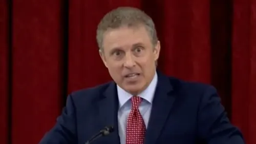 الرجل يتحدث بجدية أمام حشد، يرتدي بدلة زرقاء ورابطة عنق حمراء، مع خلفية حمراء، في سياق قانوني يتعلق بإقالته من منصبه.