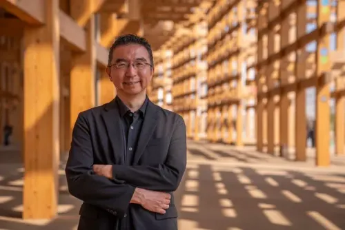 المهندس المعماري سو فوجيموتو يقف مبتسمًا داخل حلبة المعرض الخشبية الكبرى في إكسبو 2025، محاطًا بهياكل خشبية مبتكرة.