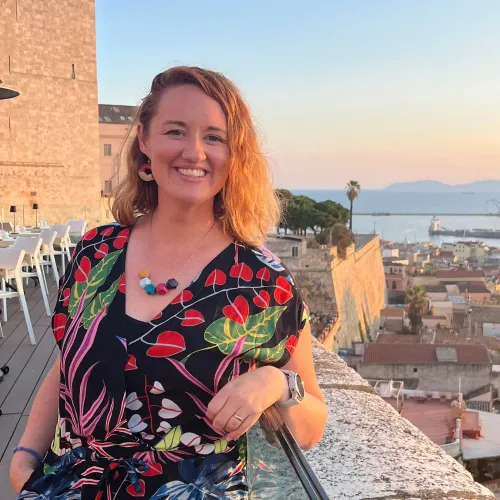 امرأة مبتسمة ترتدي ملابس ملونة، تقف في موقع مرتفع يطل على منظر خلاب لمدينة ساحلية، مع أشجار ونخيل في الخلفية.