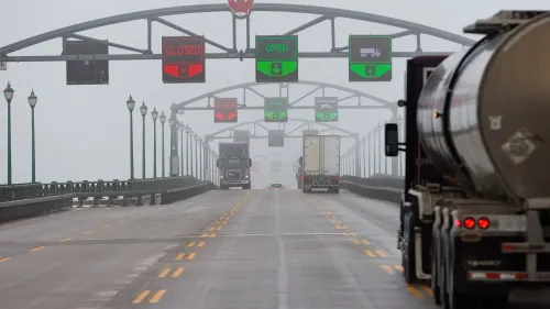 جسر يربط بين الولايات المتحدة وكندا، تظهر عليه إشارات مرورية تشير إلى حالة المرور، مع شاحنات تسير على الطريق في أجواء ضبابية.