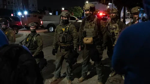 عناصر من الشرطة يرتدون زيًا عسكريًا خلال احتجاجات في لوس أنجلوس، مع وجود مركبات أمنية في الخلفية، تعكس التوترات حول مداهمات الهجرة.