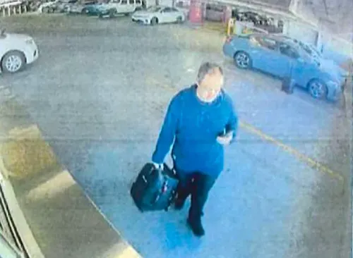 رجل يحمل حقيبة سوداء ويستخدم هاتفه، يظهر في كاميرا مراقبة داخل موقف سيارات، محاط بسيارات أخرى.