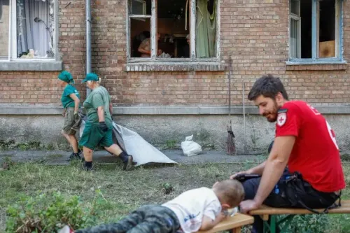 متطوعون أوكرانيون يجمعون الأنقاض من مبنى متضرر، بينما يجلس مسعف يرتدي زيًا أحمر على مقعد قريب مع طفل.