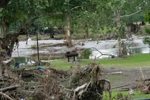 تظهر الصورة آثار الفيضانات في تكساس، حيث تتناثر الحطام والأشجار المكسورة، مع وجود مياه راكدة في الخلفية.