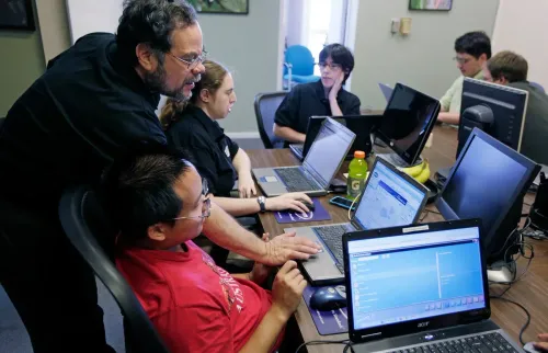 مجموعة من الأشخاص يعملون على أجهزة الكمبيوتر في بيئة تعليمية، مع وجود مدرب يوجه أحد المشاركين. يبرز التفاعل والدعم في سياق التوحد.