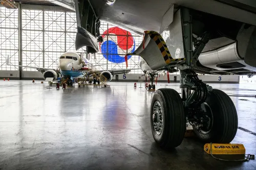 صورة لطائرة الخطوط الجوية الكورية داخل حظيرة الطائرات، مع التركيز على عجلات الطائرة والخلفية التي تظهر شعار الشركة.