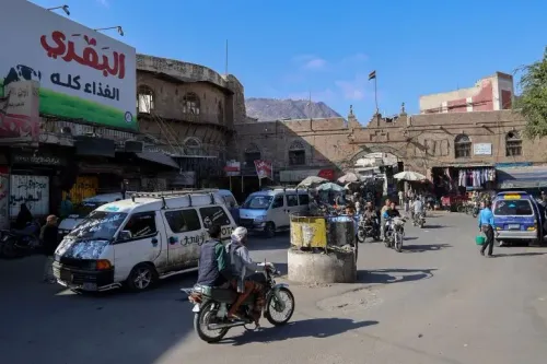 شارع مزدحم في مدينة تعز اليمنية، مع وجود سيارات ودراجات نارية، ولافتة كبيرة مكتوب عليها "النقدي الغذاء كله"، تعكس الحياة اليومية في منطقة متأثرة بالصراع.