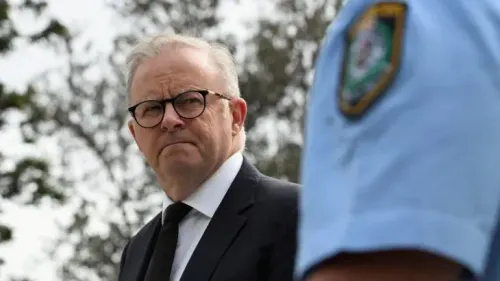 رئيس الوزراء الأسترالي أنتوني ألبانيز يتحدث بجدية خلال مؤتمر صحفي، مع وجود شرطي خلفه، في سياق إعلان عن خطة إعادة شراء الأسلحة.