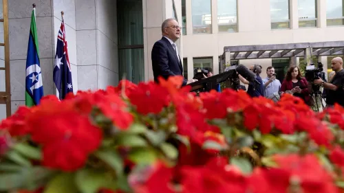 أنتوني ألبانيز يتحدث في مؤتمر صحفي للإعلان عن موعد الانتخابات الأسترالية، مع زهور حمراء تزين المشهد، خلفه أعلام أسترالية.