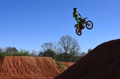 راكب دراجة نارية يقفز في الهواء خلال سباق سوبركروس، مع خلفية من الأرض الحمراء والأشجار. تعكس الصورة الإثارة والتحدي في هذه الرياضة الخطيرة.