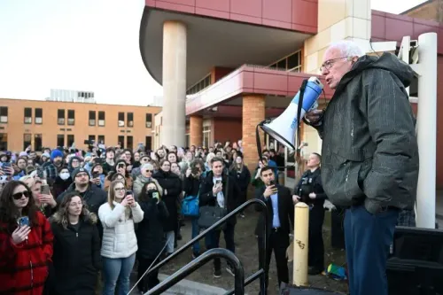 ساندرز يتحدث إلى حشد كبير من المؤيدين باستخدام ميكروفون، مع وجود علامات احتجاج في الخلفية، خلال جولة لمواجهة إدارة ترامب.