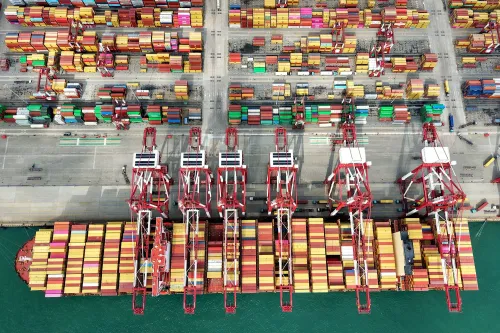 صورة جوية لميناء يحتوي على حاويات ملونة متعددة، مع رافعات تحميل كبيرة، تعكس حركة التجارة العالمية وتأثير التعريفات الجمركية.