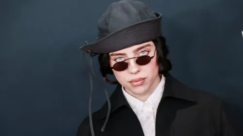 بيلي إيليش ترتدي نظارات شمسية وقبعة، تعبيرها جاد، في خلفية داكنة. تعكس الصورة أسلوبها الفريد وثقتها في نفسها.