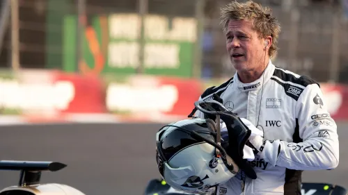 براد بيت في زي سائق سيارات سباق، يحمل خوذته بعد انتهاء جولة في حلبة السباق، مع خلفية لافتة تحمل شعار سباق.