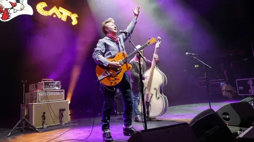 براين سيتزر يؤدي على المسرح مع فرقة Stray Cats، مع إضاءة ملونة وأجواء حماسية، رغم معاناته من مشكلة صحية تؤثر على قدرته على العزف.