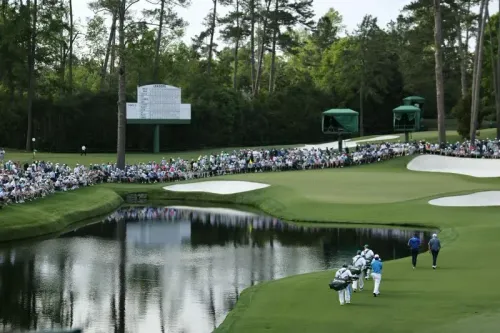 جمهور كبير يشاهد لاعبي الغولف يتنافسون في ملعب أوغوستا ناشيونال، مع وجود لوحة النتائج والمياه في الخلفية.