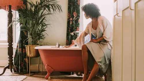 امرأة تجلس بجانب حوض استحمام وردي، تتفاعل مع الماء في بيئة مريحة، تعكس أهمية العناية الذاتية والتحفيز الذاتي.