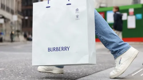 حقيبة تسوق بيضاء تحمل شعار بربري، تُظهر شخصًا يرتدي حذاءً رياضيًا، في إطار حضري يعكس التحديات التي تواجه العلامة التجارية.