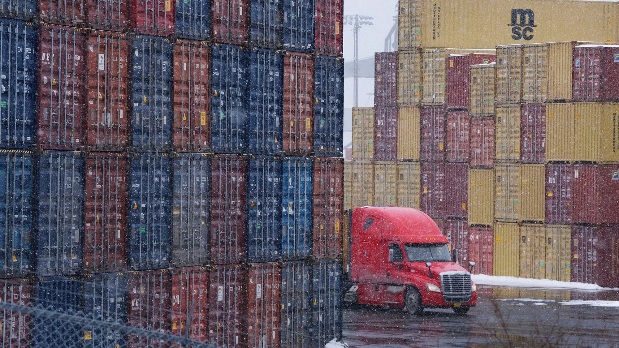 شاحنة حمراء تمر بين حاويات شحن ملونة في ميناء، تعكس تأثير سياسات ترامب التجارية على الشركات الصغيرة والاقتصاد الأمريكي.