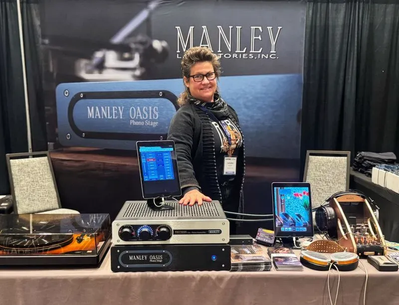 امرأة تقف أمام طاولة تعرض معدات صوتية من شركة Manley Labs، مع خلفية تحمل شعار الشركة، تعكس تأثير التعريفات الجمركية على الأعمال الصغيرة.