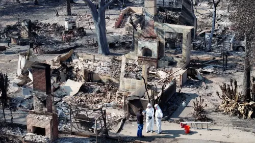 موقع حريق مدمر في كاليفورنيا، يظهر بقايا المنازل المحترقة، مع أشخاص يرتدون ملابس واقية يتحدثون في الموقع.