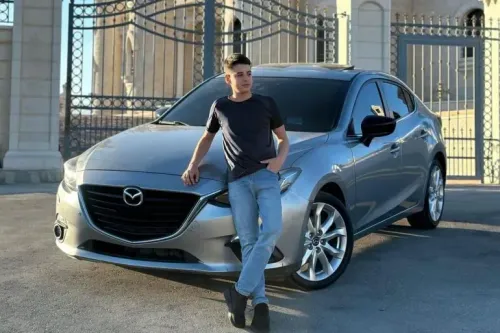 محمد إبراهيم، مراهق فلسطيني أمريكي، يقف بجانب سيارة فضية أمام بوابة، مع خلفية معمارية. الصورة تعكس قضيته المعقدة في السجون الإسرائيلية.