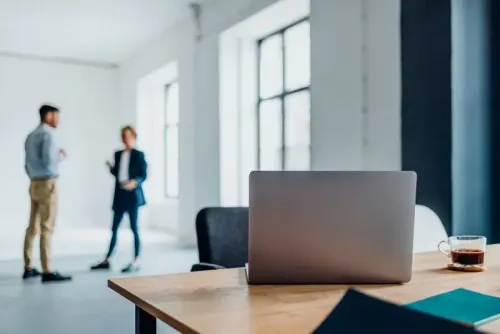 مكتب حديث يظهر جهاز كمبيوتر محمول على الطاولة مع فنجان قهوة، بينما يتحدث شخصان في الخلفية حول موضوعات العمل.