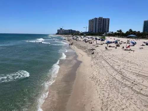 شاطئ مشمس في ميامي مع زوار يستمتعون بالرمال والماء، بينما تظهر المباني الشاهقة في الخلفية، تعكس تزايد اهتمام الكنديين بالعقارات الأمريكية.