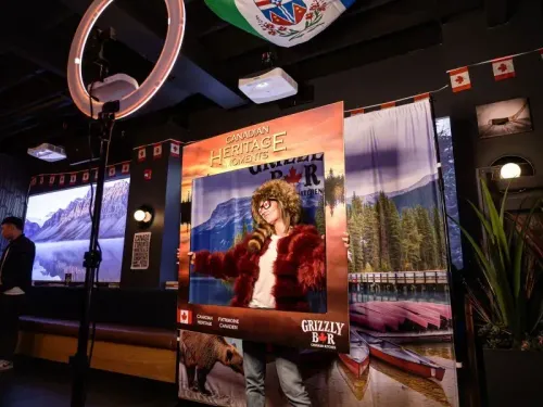 امرأة ترتدي زيًا كنديًا تقف داخل إطار يحمل شعار "لحظات التراث الكندي" في مطعم، مع صور طبيعية خلفها، تعكس الثقافة الكندية.