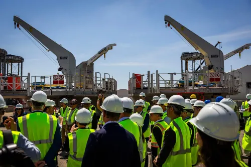 مجموعة من العمال مع معدات بناء في موقع مشروع طاقة الرياح "Revolution Wind" في رود آيلاند، مع التركيز على الآلات المستخدمة في المشروع.