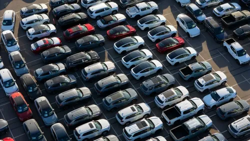 مصفوفة من السيارات الجديدة في موقف سيارات، تشمل ألوانًا متنوعة مثل الأبيض والأسود والأحمر، تعكس السوق الأمريكي وتأثير التعريفات الجمركية.