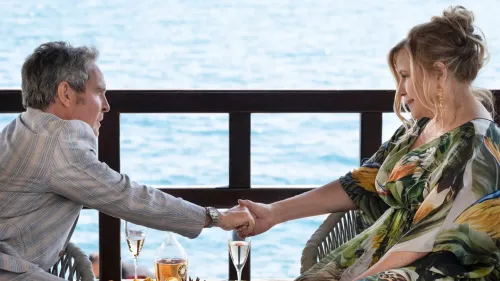 مشهد يظهر شخصين يجلسان على شرفة تطل على البحر، يتبادلان الأيدي بلمسة حميمية، مع مشروبات أمامهما، مما يعكس أجواء درامية من مسلسل "اللوتس الأبيض".