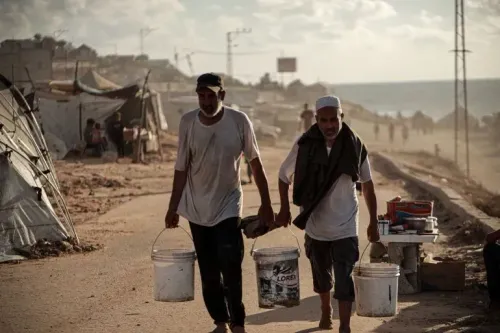 رجلان في مخيم للاجئين بدير البلح، يحملان دلوين، بينما تظهر خيام ومشهد رملي في الخلفية، تعبيرًا عن الحياة اليومية في ظل النزاع.