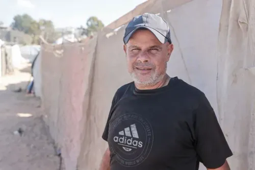 رجل مبتسم يقف أمام خيام في مخيم للاجئين في دير البلح، غزة، معبرًا عن الأمل وسط الأوضاع الصعبة بعد إعلان خطة ترامب لوقف إطلاق النار.
