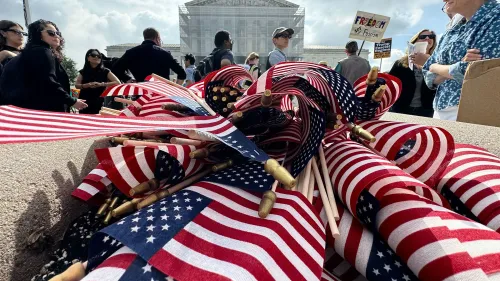 تتراكم أعلام أمريكية صغيرة تحمل ألوان العلم، مع وجود متظاهرين في الخلفية يحملون لافتات تعبر عن آرائهم بشأن سياسة ترامب.