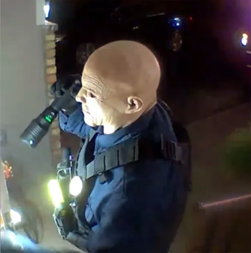 شخص يرتدي زي ضابط شرطة ويحمل مصباحًا، يظهر في مشهد مشبوه، مما يعكس تصاعد العنف السياسي في الولايات المتحدة.